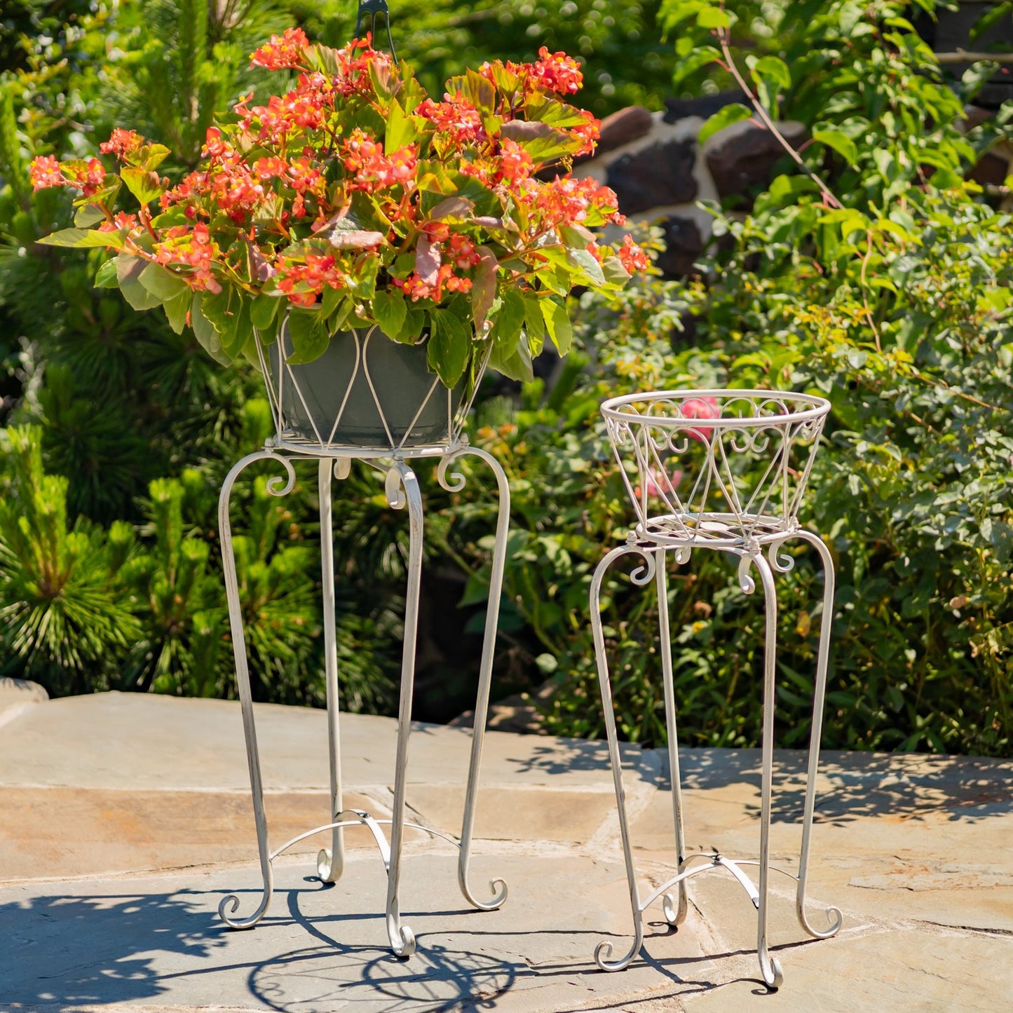 Two white metal garden plant stands outdoors on a stone patio, with one holding a green planter overflowing with orange flowers and the other empty.