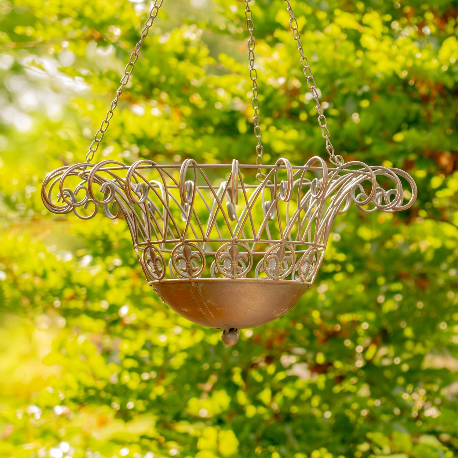 A hanging metal planter basket with ornate scrollwork and a shallow bowl, suspended by chains against a bright green garden background.
