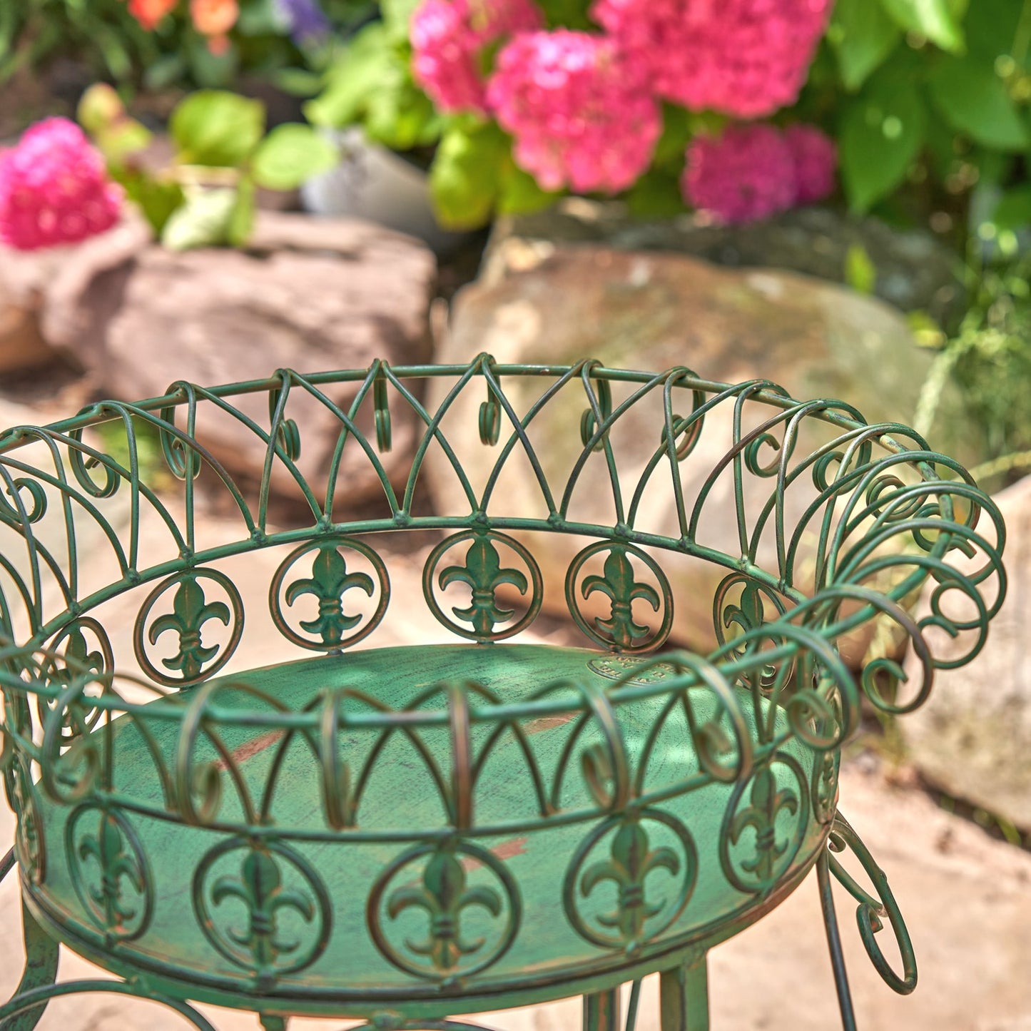 A green metal garden planter stand with an ornate scrollwork design and fleur-de-lis motifs, shown outdoors in a garden setting with blurred pink flowers and rocks in the background.