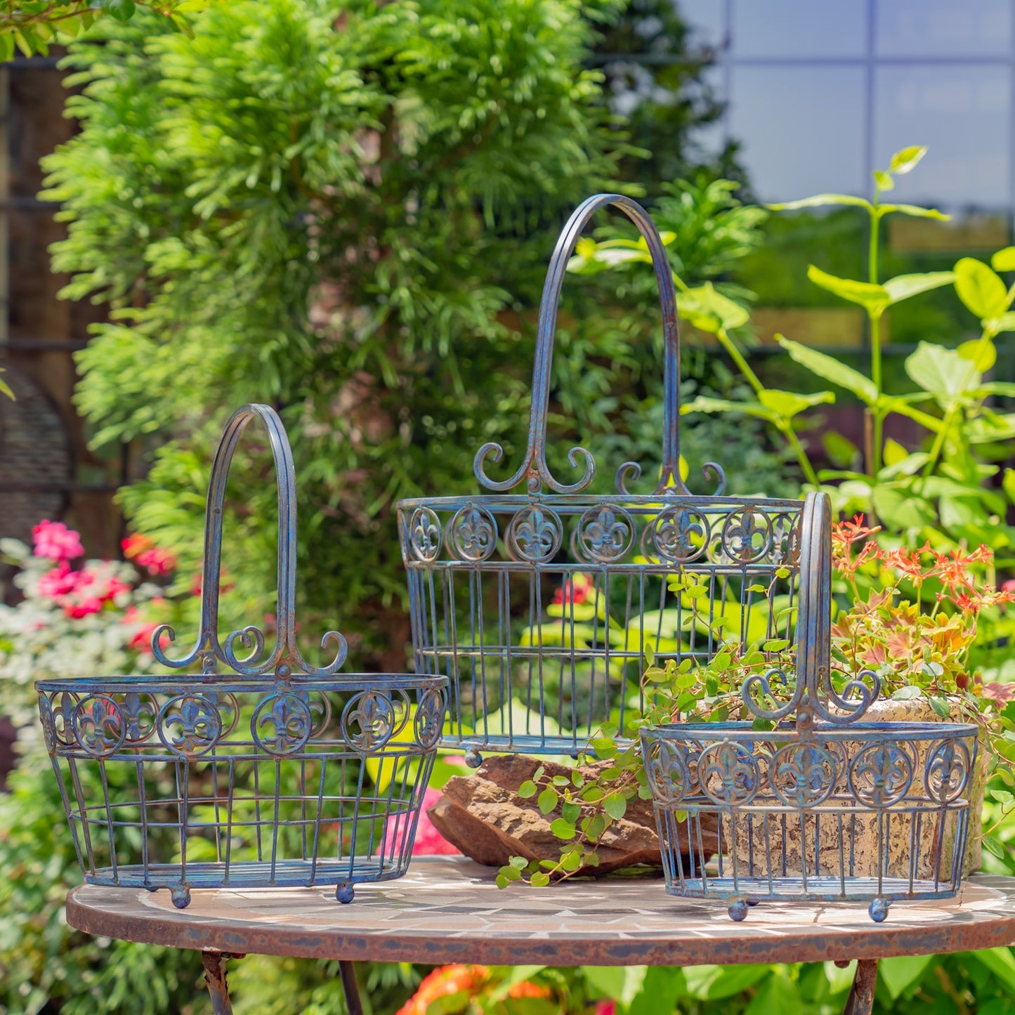 Three decorative metal plant baskets in a distressed blue finish, displayed on a rustic wooden table in a lush garden setting.