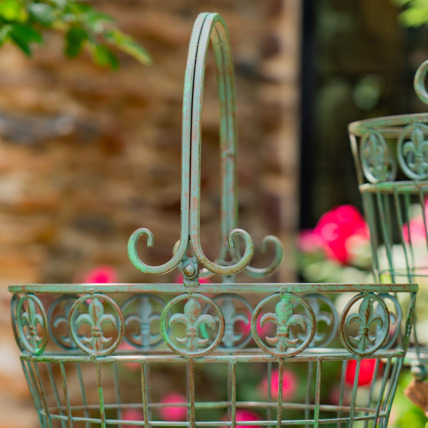 A close-up of an ornate metal garden basket with a hinged top handle and decorative circular cutouts, finished in a weathered green patina. The background is a softly blurred outdoor garden scene with a brick wall and pink flowers.