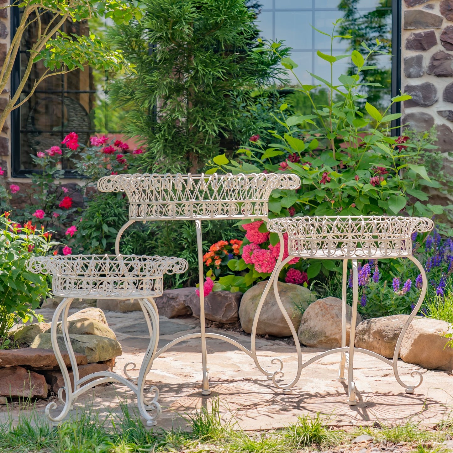 Three white wrought-iron garden tables of different sizes arranged on a stone patio in a lush garden setting with flowers and greenery.