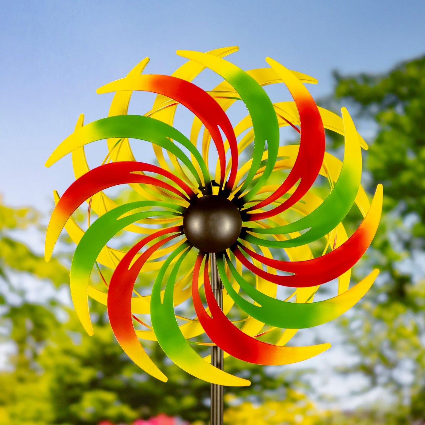 A colorful metal garden wind spinner with alternating red, green, and yellow curved blades, mounted on a black central hub and a thin metal stake, set against a blurred outdoor background of green trees and blue sky.