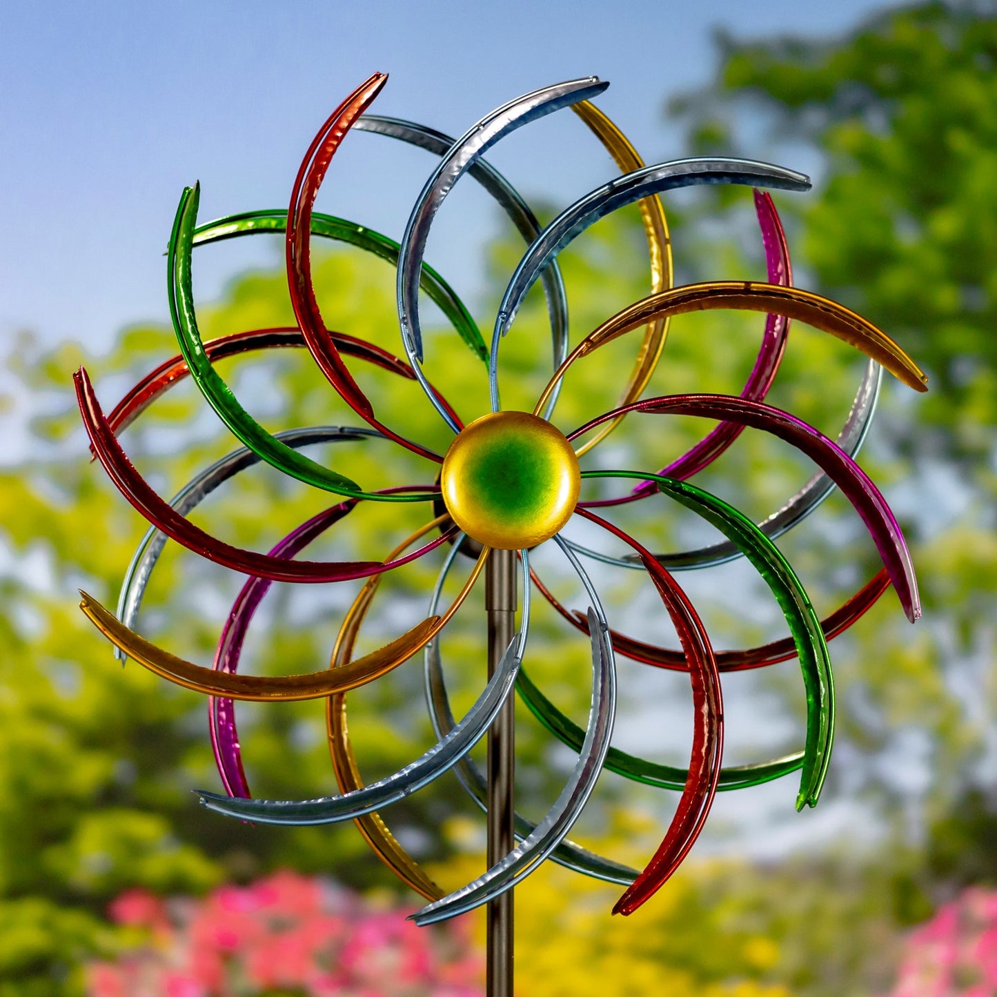 A colorful metal wind spinner garden stake with multiple curved blades in red, green, purple, gold, and silver, set against a blurred background of green foliage and blue sky.