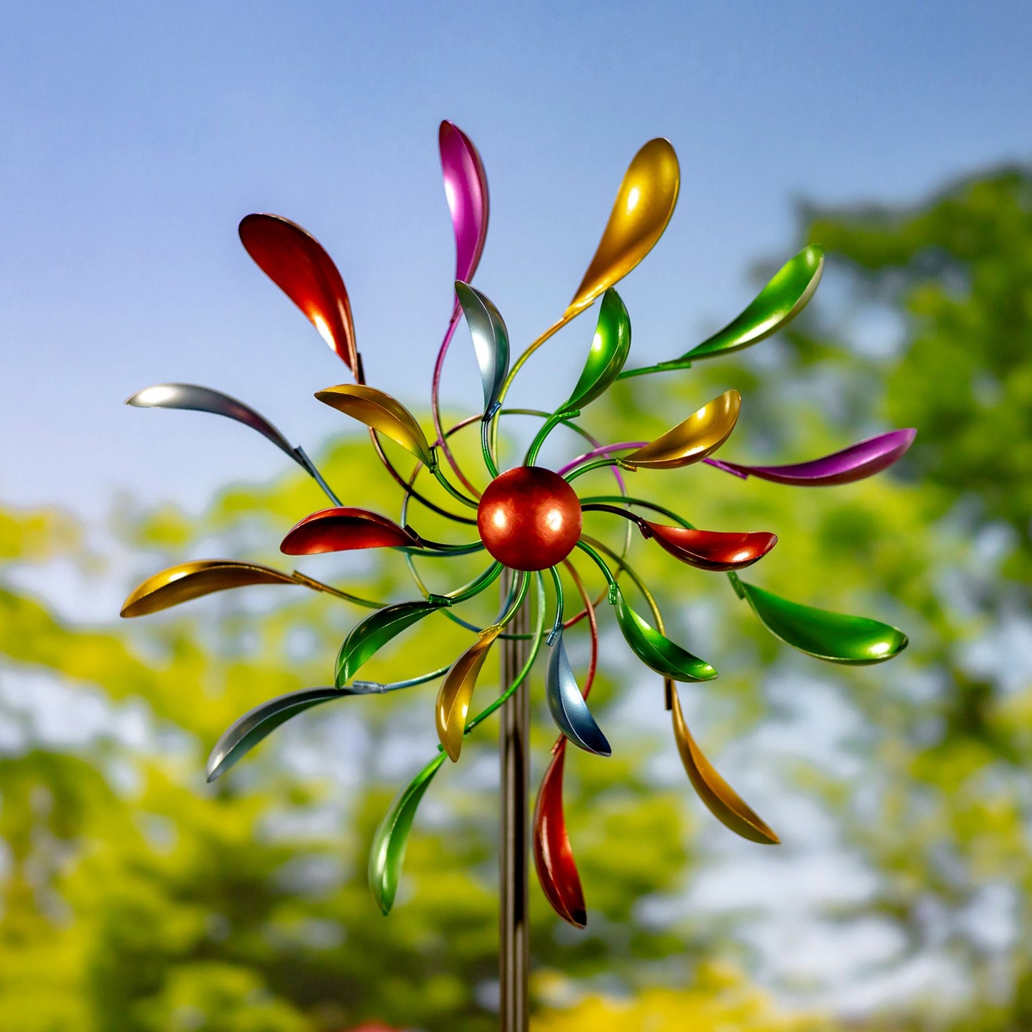 A colorful metal garden wind spinner with a red central sphere and multiple iridescent leaf-shaped blades in green, gold, red, purple, and silver, mounted on a thin metal stake against a blurred outdoor background of blue sky and green foliage.