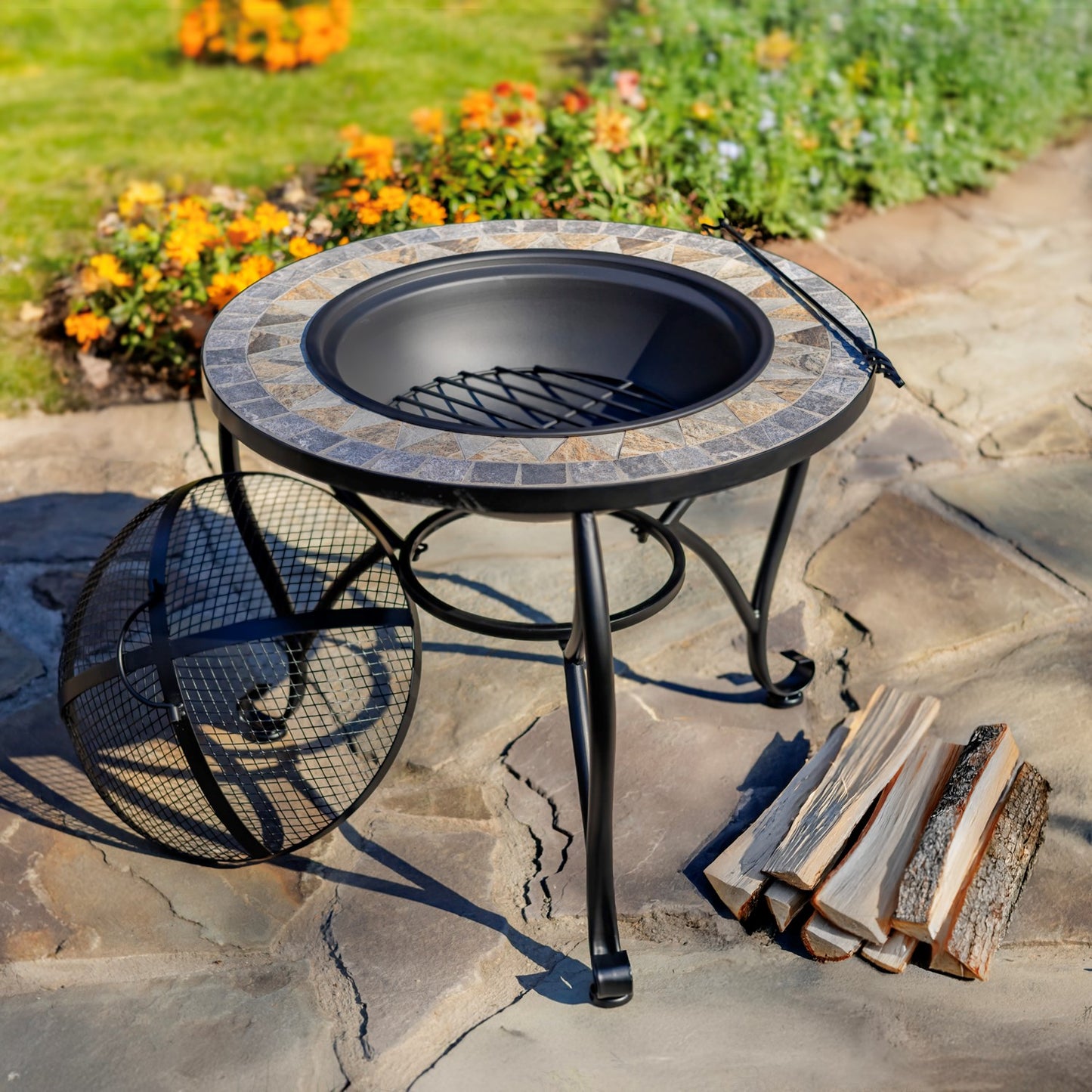 Outdoor round fire pit table with a black metal frame and a stone-tile patterned rim, shown on a stone patio with firewood and a garden bed in the background.