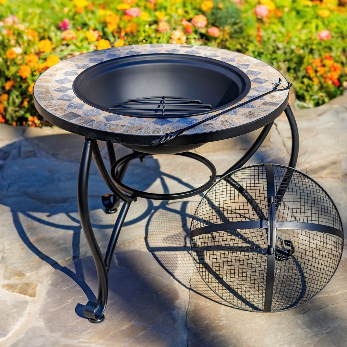 Outdoor round fire pit table with a mosaic tile-patterned rim and black metal legs, shown on a stone patio with flowers in the background. A matching round fire screen is placed beside the table.