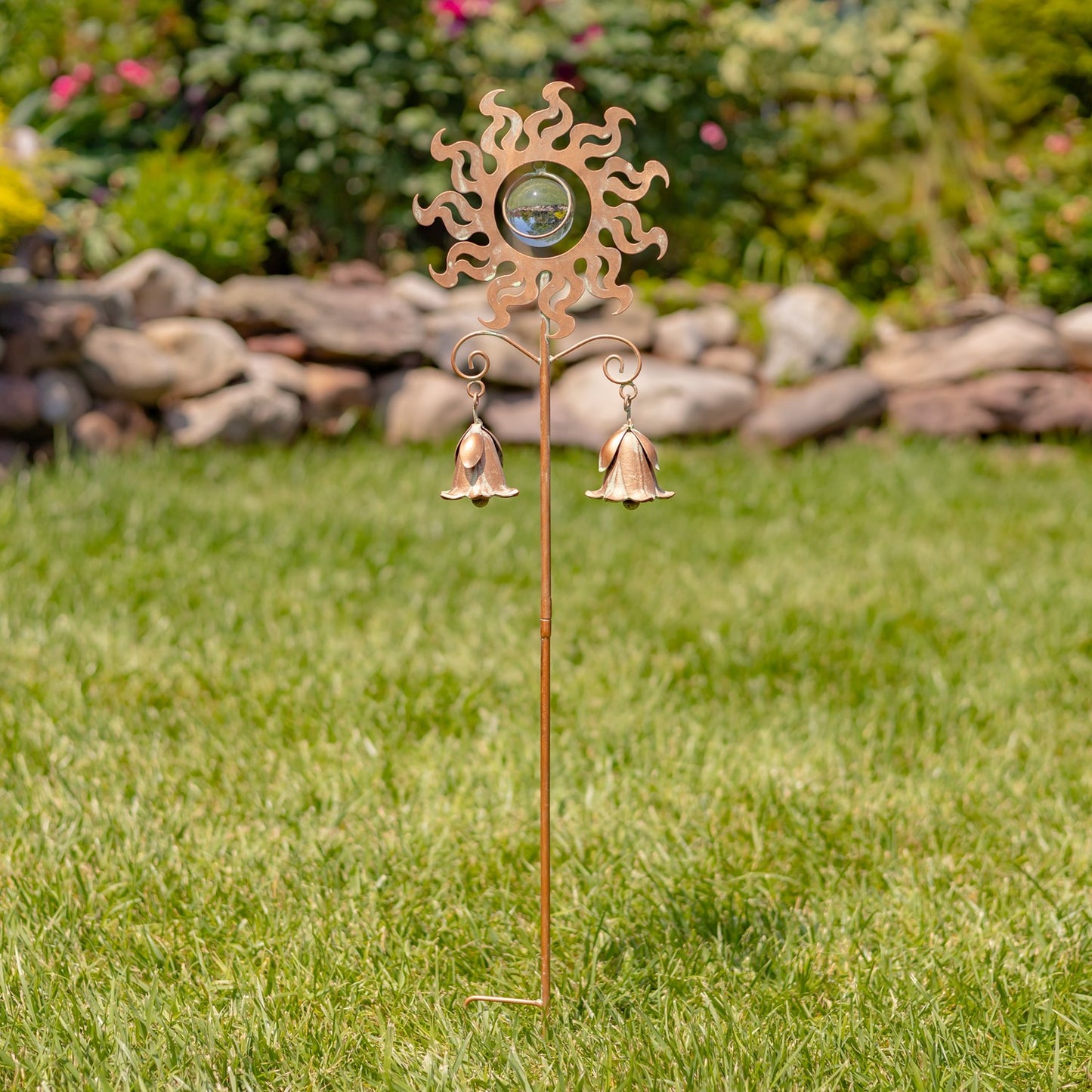 A decorative metal garden stake with a sunburst top, a clear glass bubble in the center, and two small hanging bells, photographed outdoors on a green lawn with a blurred garden background.