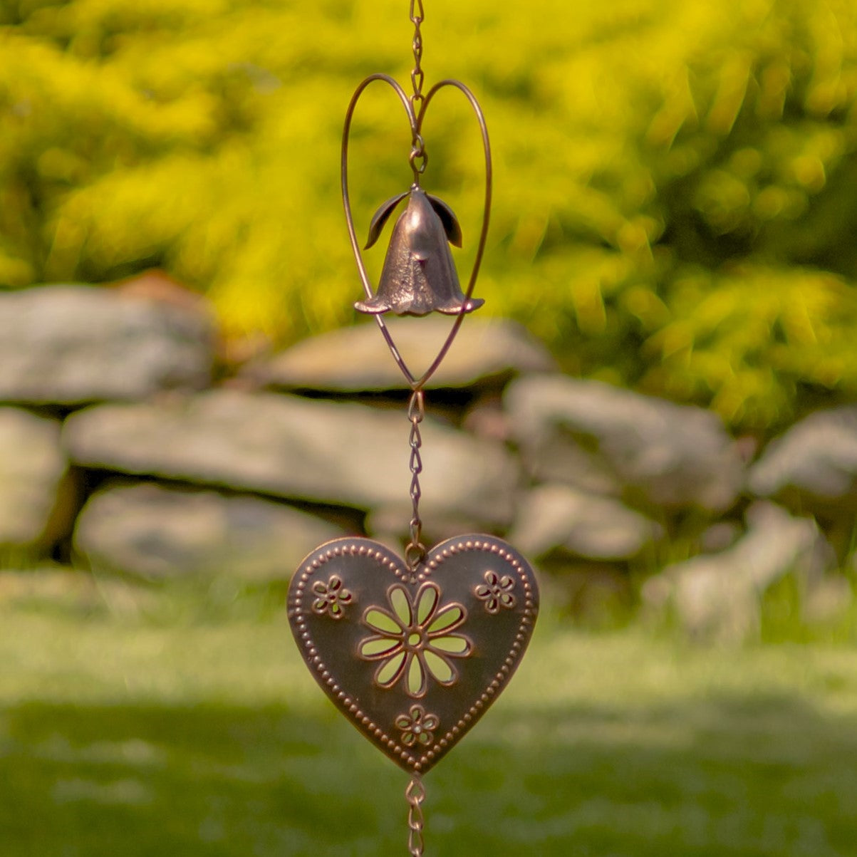 A copper-toned metal garden wind spinner featuring a bell-shaped flower suspended inside a heart-shaped wire frame, set against a blurred green grass background.