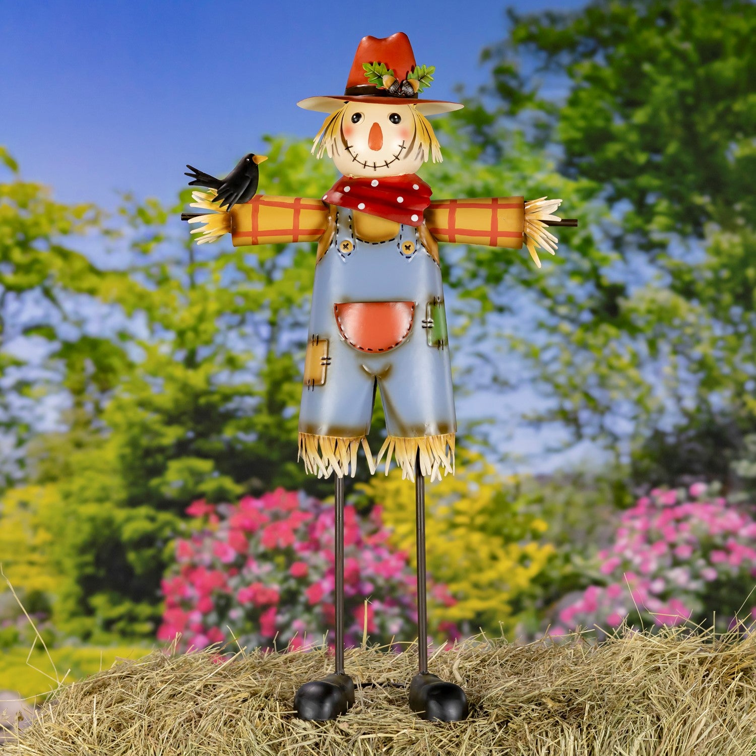 A metal garden scarecrow decoration standing on a hay bale with a blurred outdoor garden background. The scarecrow features a straw hat with green leaves, a red polka-dot neck scarf, a blue body, yellow straw arms and hair, and a small black bird perched on its left arm.