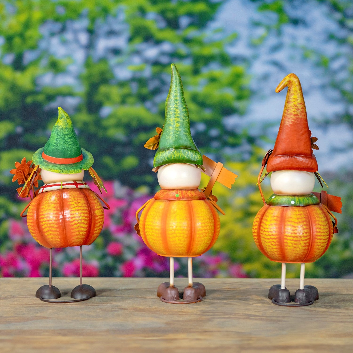 Three small decorative gnome figures shaped like pumpkins, standing on a wooden surface with a blurred green garden background. Each gnome has a pointed hat, a white head, and orange-yellow pumpkin bodies.