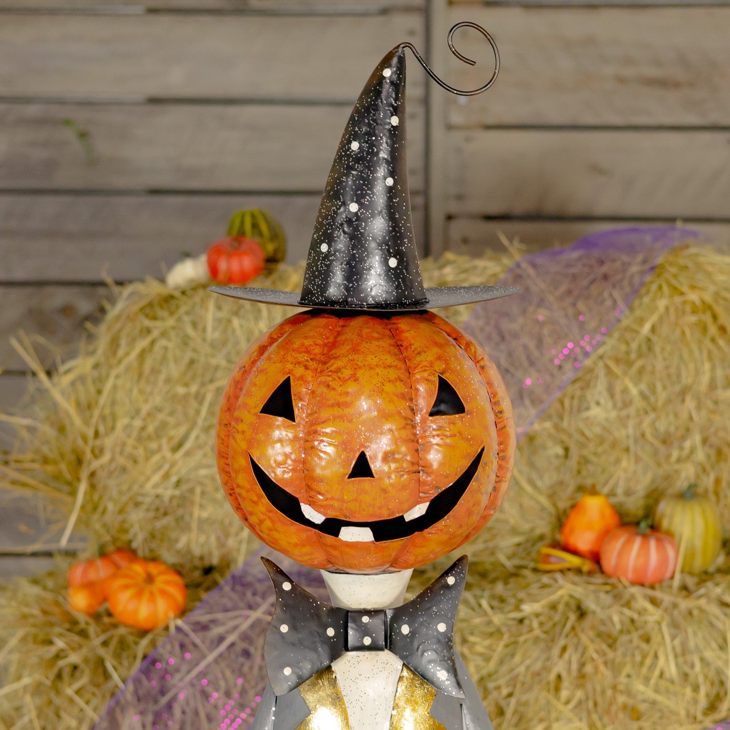 A decorative Halloween pumpkin figurine with a carved jack-o'-lantern face, wearing a black witch hat with white polka dots and a black bow tie with white dots, standing on hay bales with small pumpkins and a rustic wood backdrop.