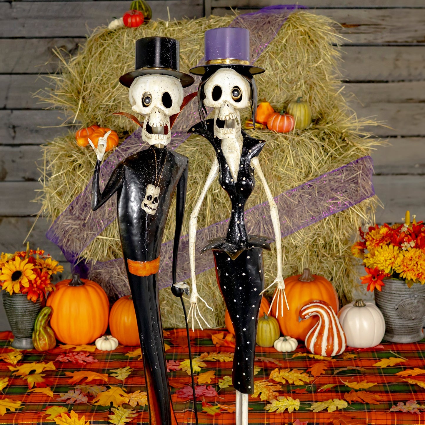 Two Halloween skeleton figurines dressed in black outfits, standing on a plaid autumn tablecloth with pumpkins, flowers, and a hay bale backdrop.