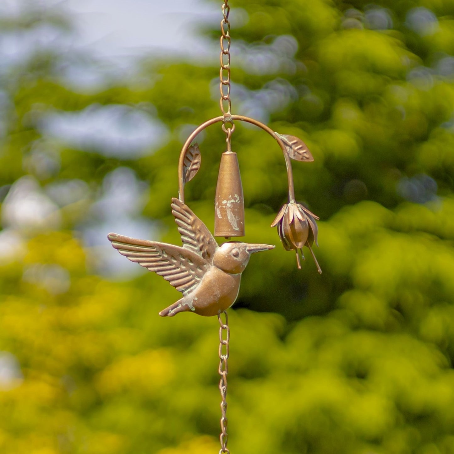 A bronze metal garden wind chime featuring a hummingbird and a bell-shaped flower, suspended by a chain against a blurred green foliage background.