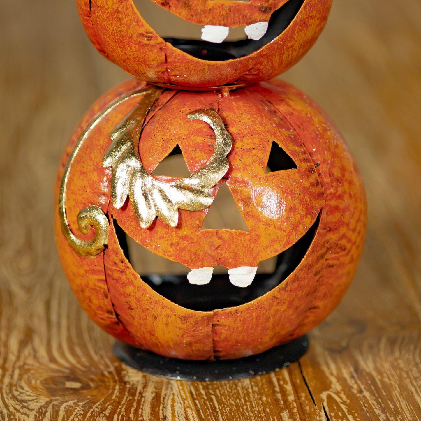 A stacked Halloween jack-o'-lantern decoration made from two overlapping orange pumpkins, with one pumpkin on top partially visible and the lower pumpkin featuring a carved face, white teeth, and a metallic gold scroll accent on the side, set on a wooden surface.