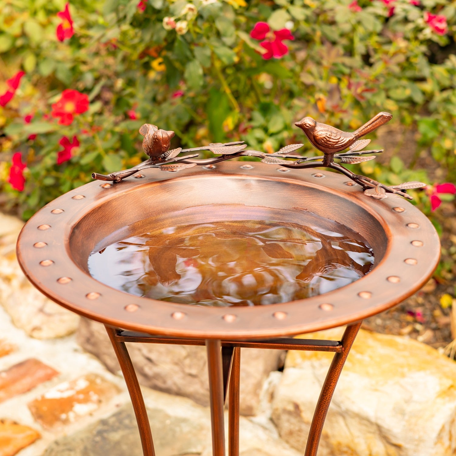 A copper bird bath with a matching stand, featuring decorative bird sculptures on the rim, set in a garden with flowers and rocks in the background.