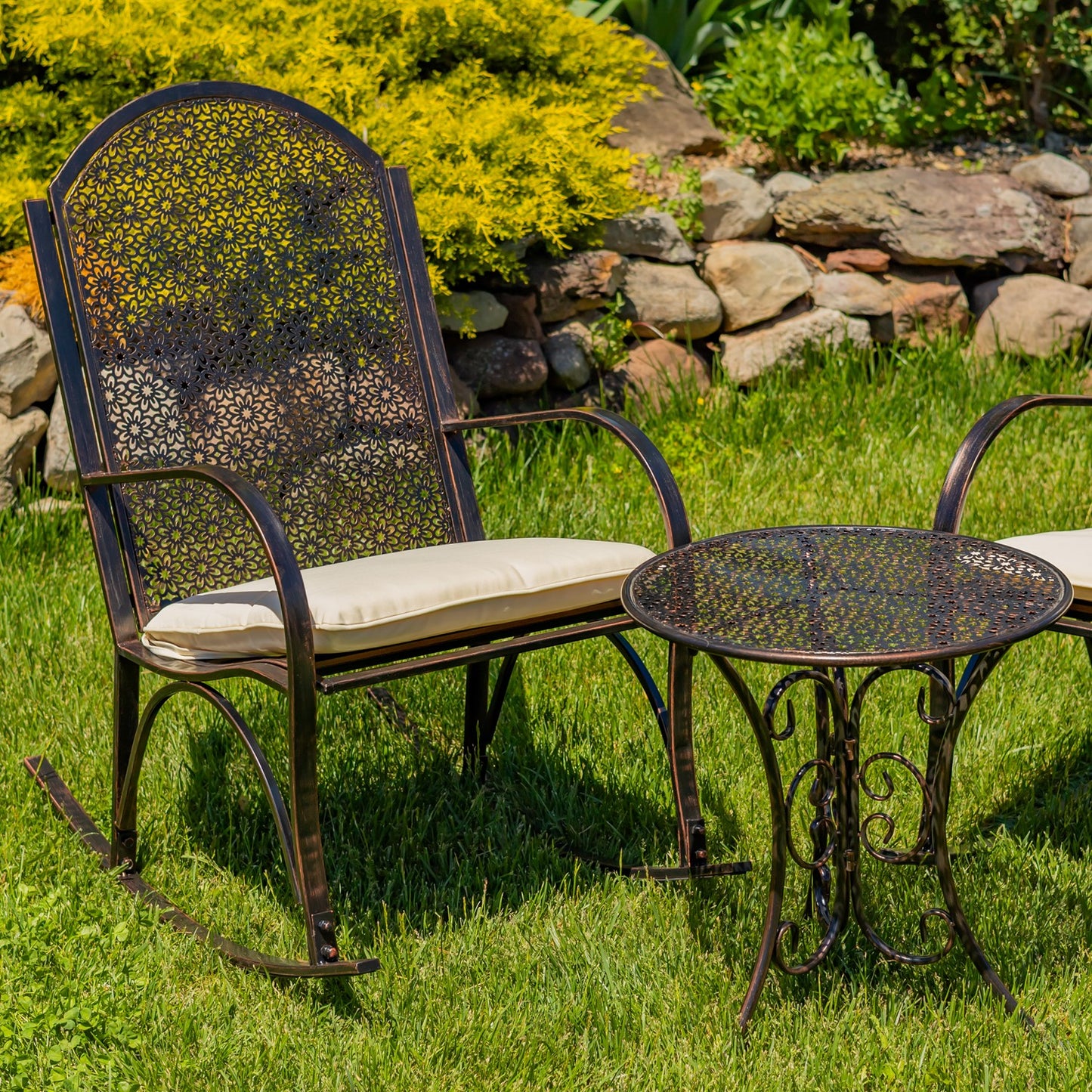 Outdoor patio furniture set on a grassy lawn, featuring a dark metal rocking chair with a cream cushion and a matching round bistro table, with a stone garden wall and yellow-green shrubs in the background.