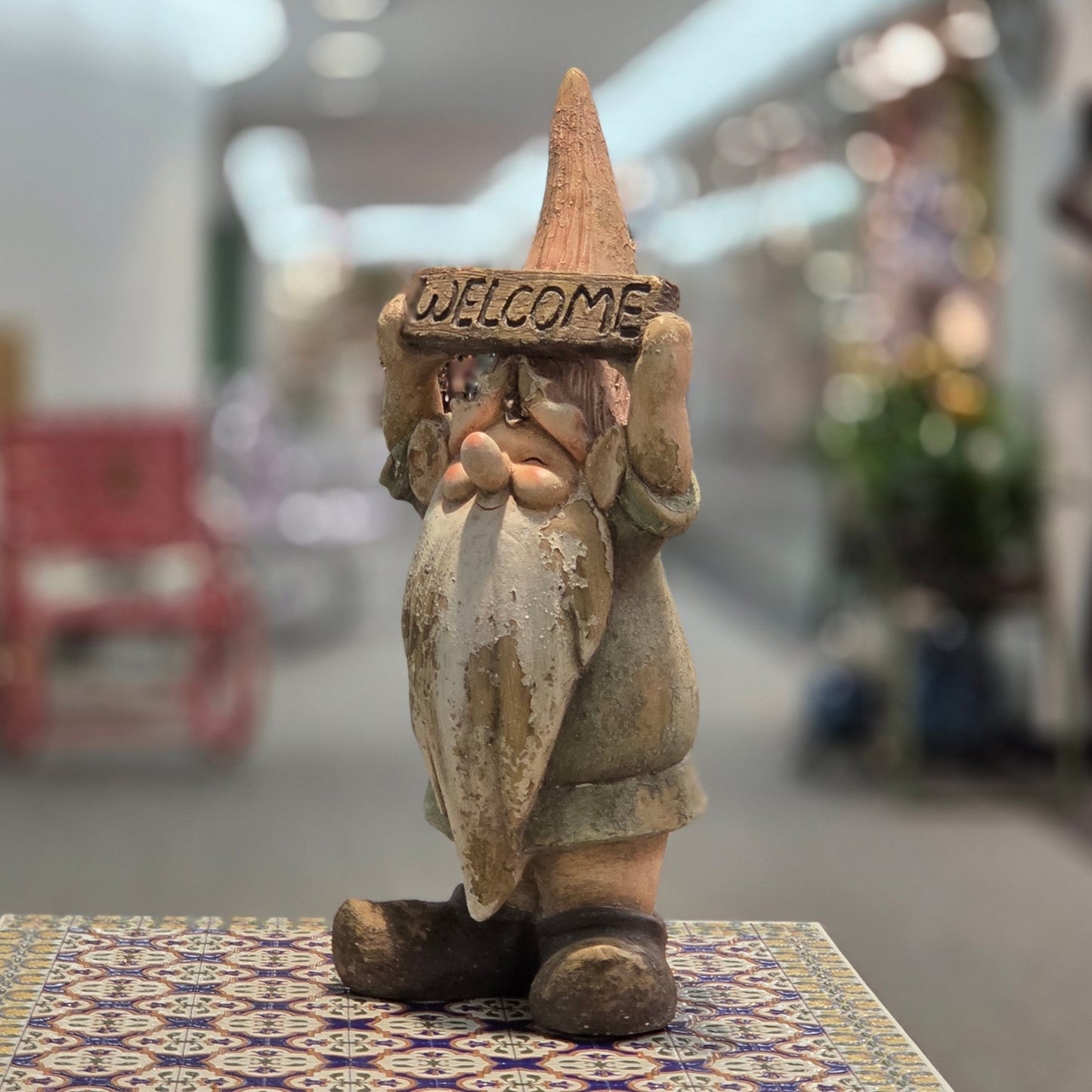 A rustic, hand-painted clay gnome figurine holding a 'WELCOME' sign, displayed on a patterned tile surface with a blurred indoor background.