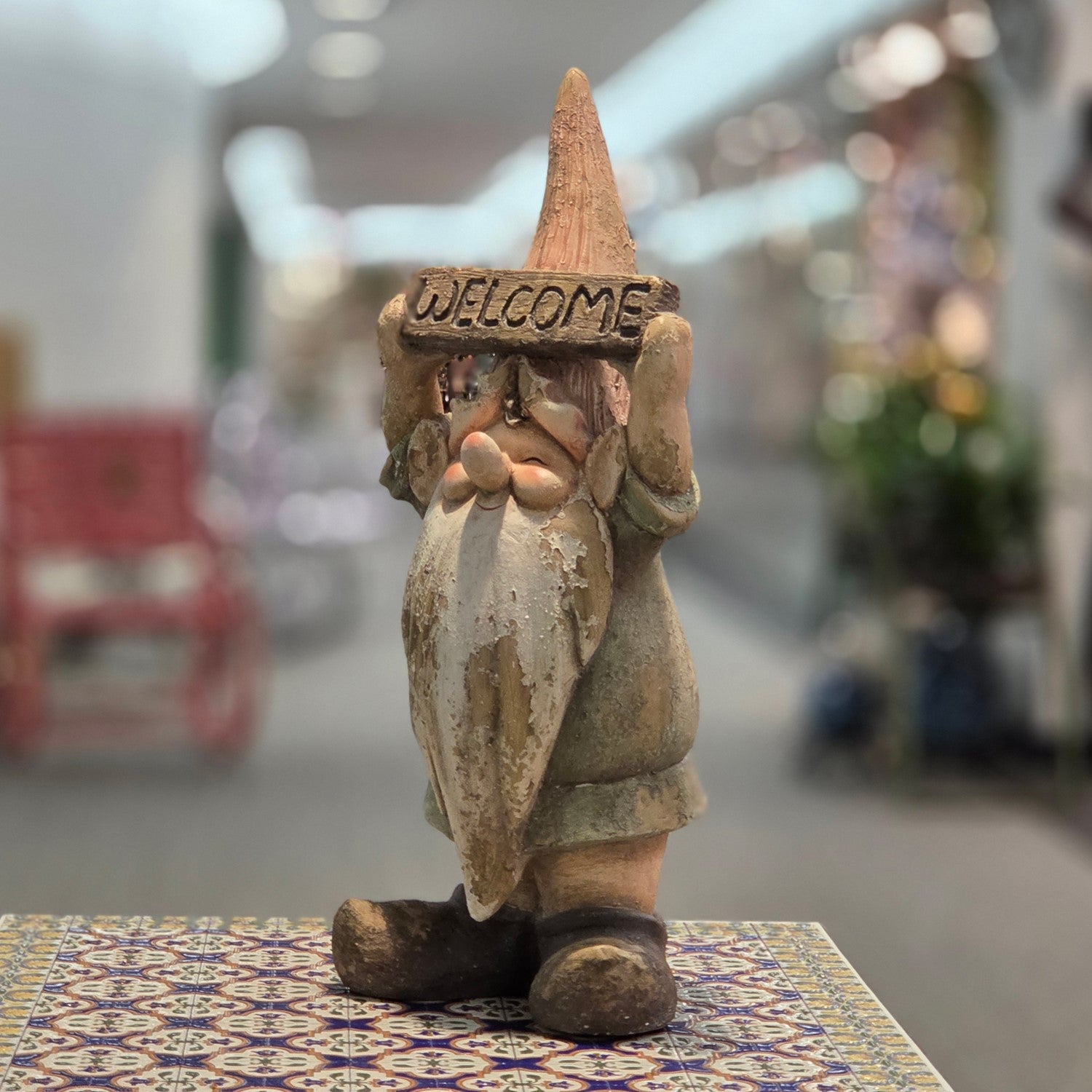 A rustic, hand-painted clay gnome figurine holding a 'WELCOME' sign, displayed on a patterned tile surface with a blurred indoor background.