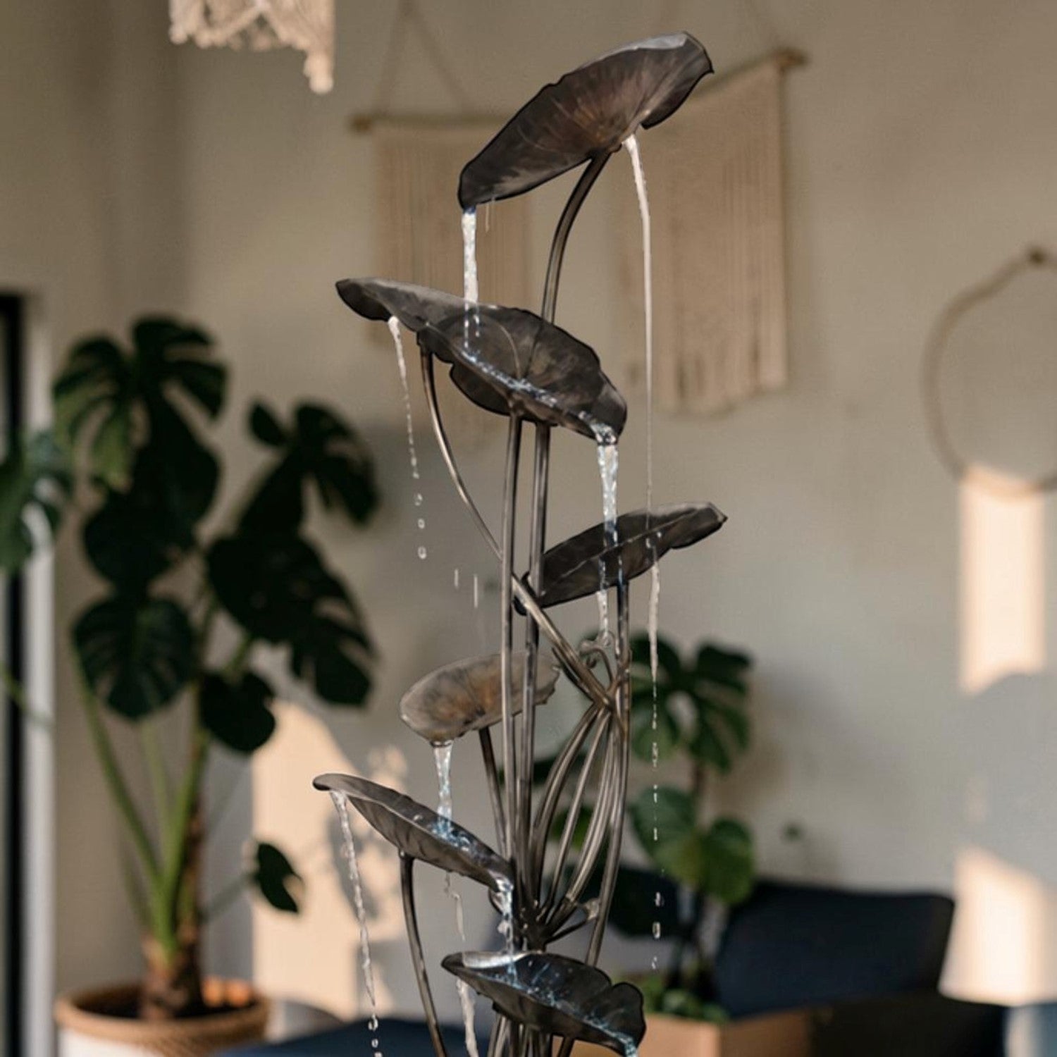 A metal waterfall fountain with leaf-shaped plates and flowing water, styled in a modern living room setting with plants and home decor in the background.