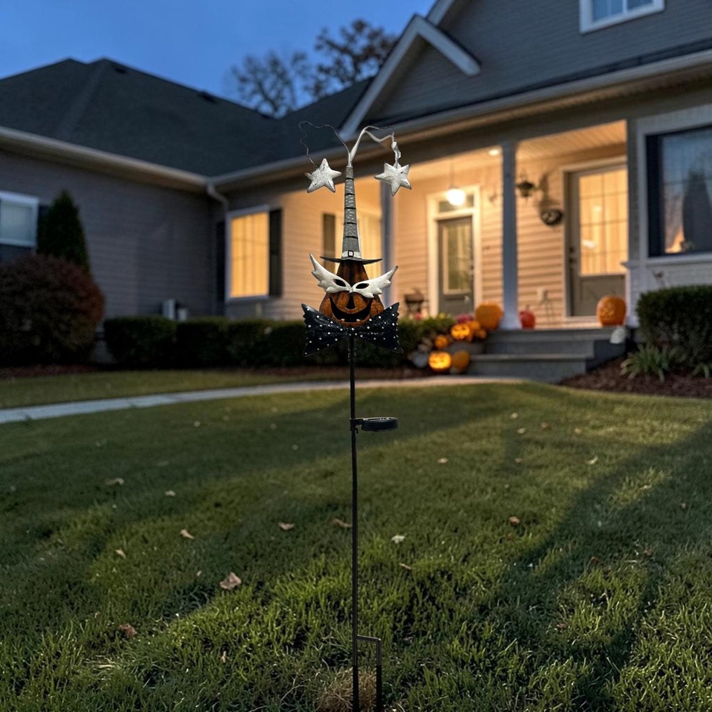 A Halloween yard decoration featuring a witch's hat with stars and a jack-o'-lantern, mounted on a stake in a front yard at dusk.