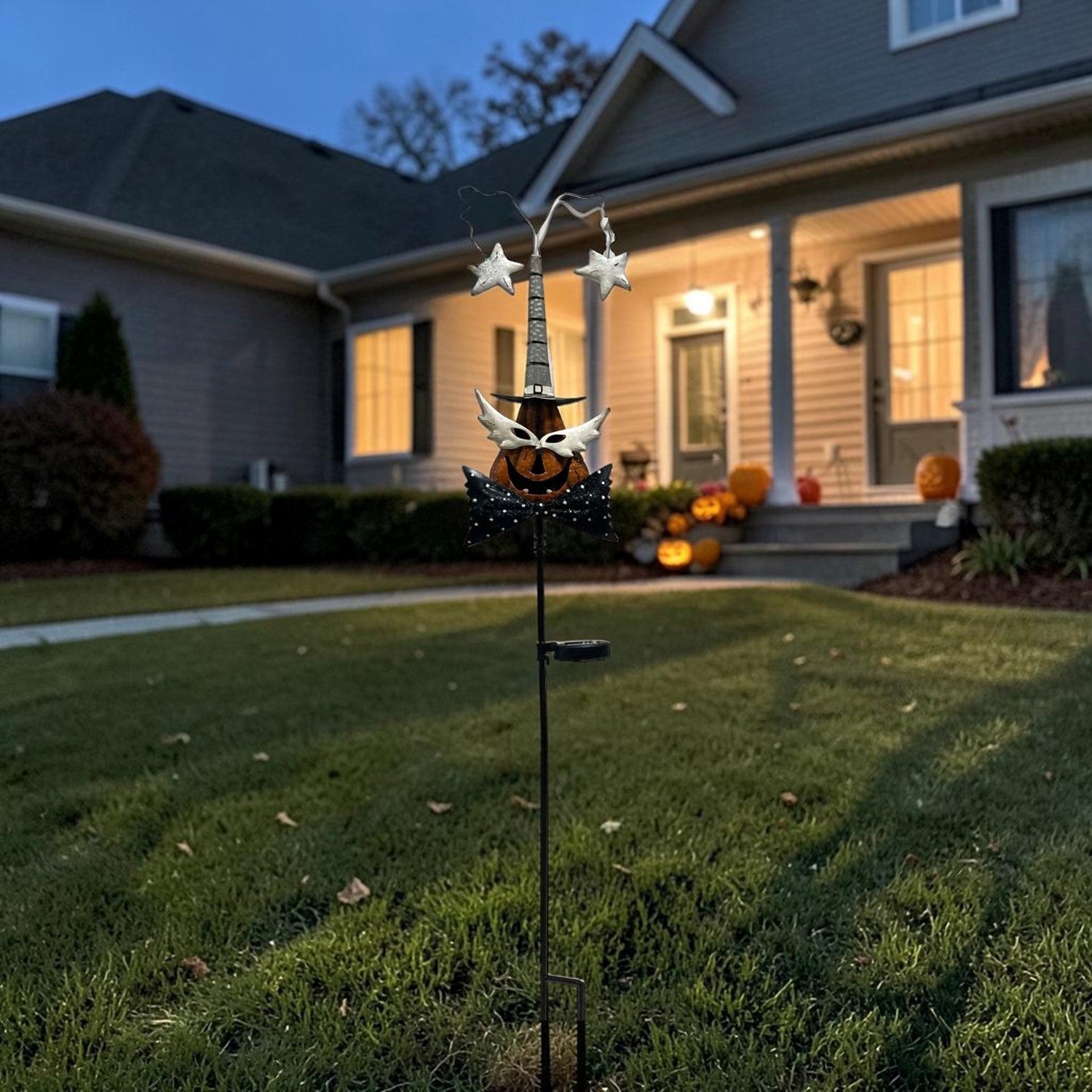 A Halloween yard decoration featuring a witch's hat with stars and a jack-o'-lantern, mounted on a stake in a front yard at dusk.