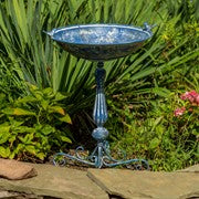 A blue metal garden bird bath with a pedestal stand, set outdoors in a garden with green plants and a stone path.
