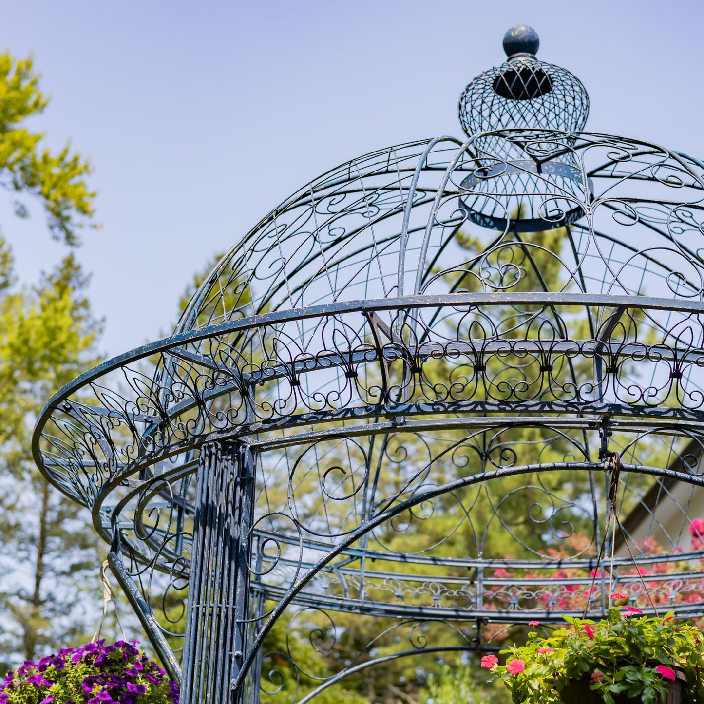 An ornate metal garden gazebo with a domed roof and decorative scrollwork, photographed outdoors against a blue sky and green trees.