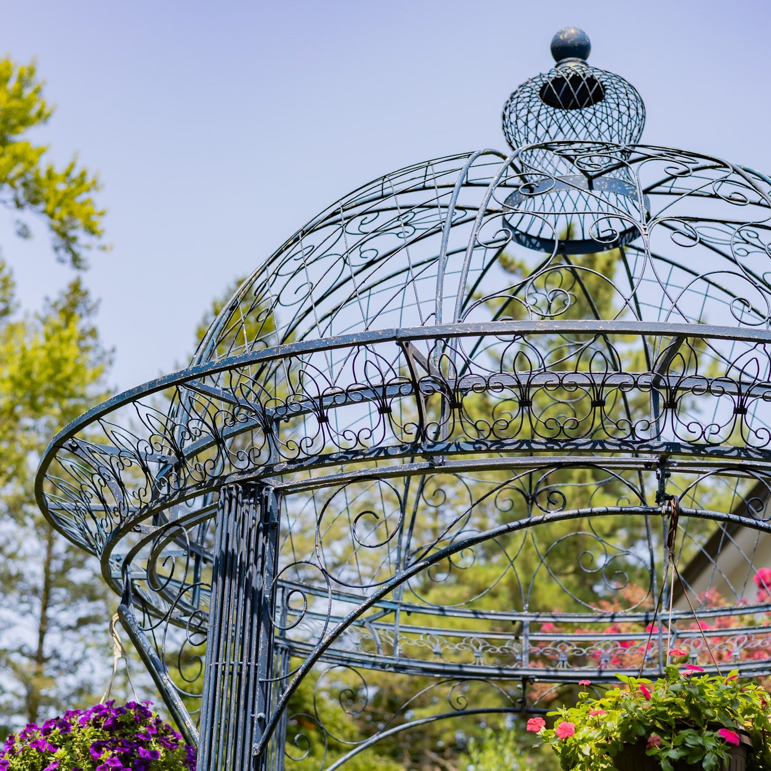 An ornate metal garden gazebo with a domed roof and decorative scrollwork, photographed outdoors against a blue sky and green trees.