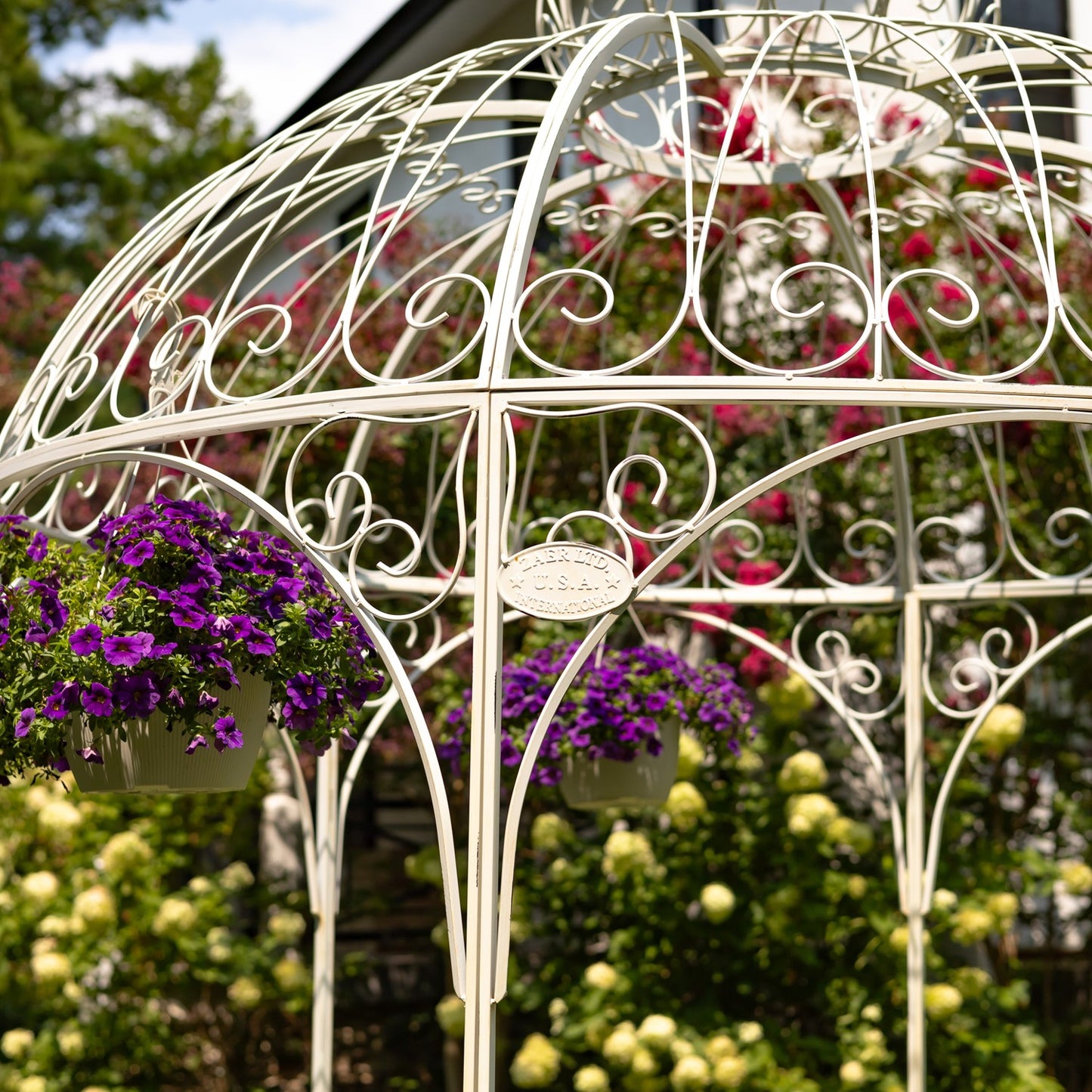 A white ornate metal garden gazebo or trellis with hanging purple petunia flower baskets, set in a lush garden with green foliage and pink flowers in the background.