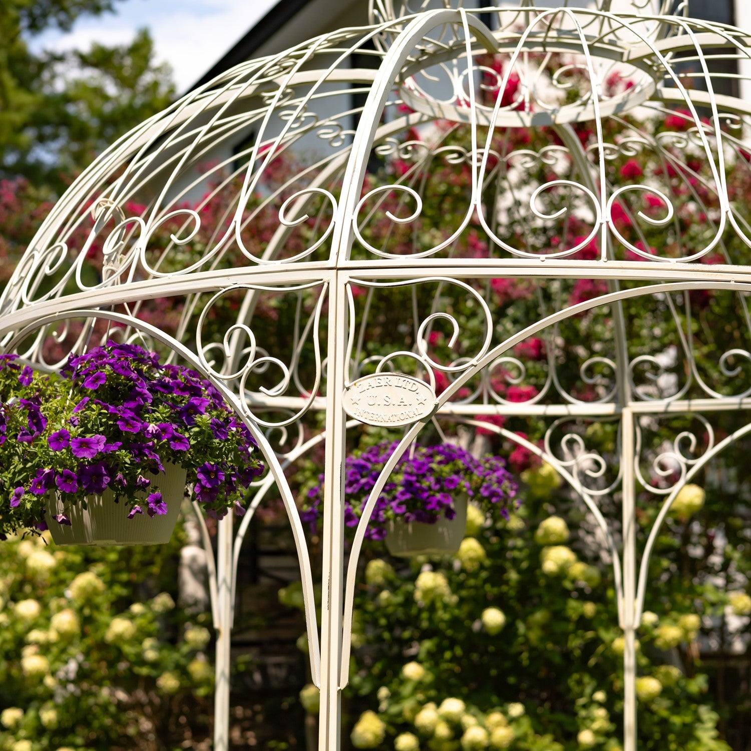 A white ornate metal garden gazebo or trellis with hanging purple petunia flower baskets, set in a lush garden with green foliage and pink flowers in the background.