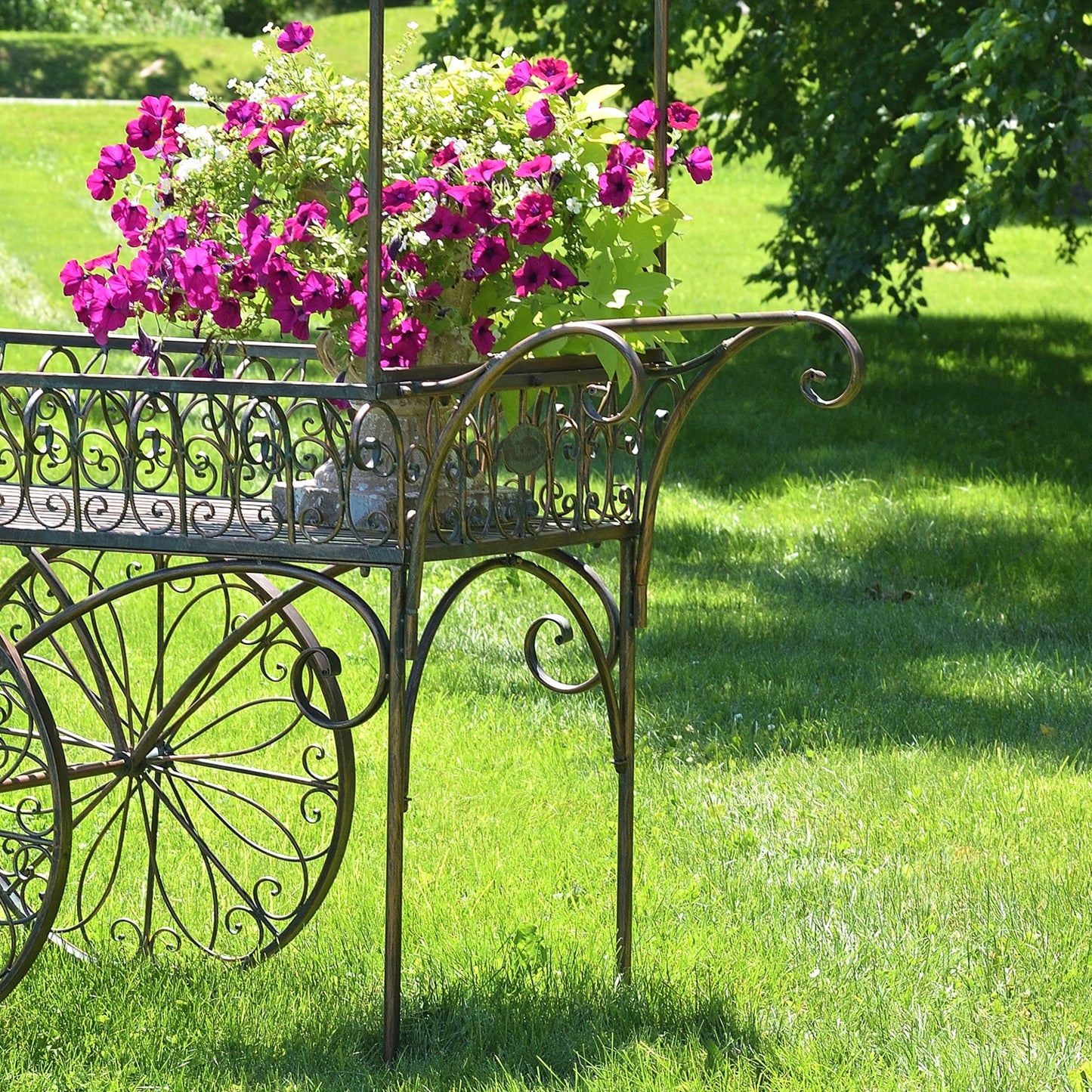 An ornate metal garden cart with decorative scrollwork and large wheels, placed on a green lawn and holding a large vase of bright pink flowers.