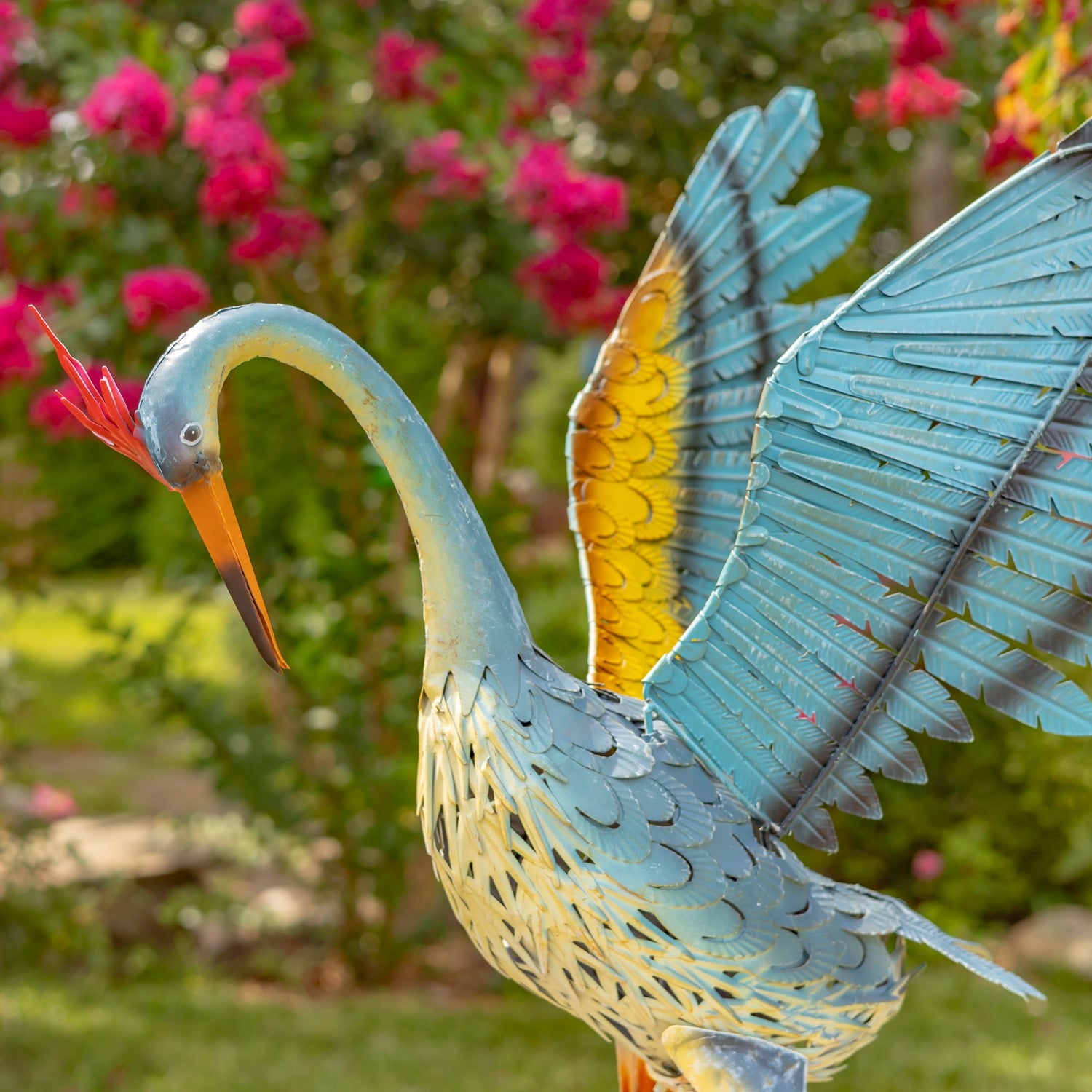 A decorative metal garden statue of a heron with spread wings, featuring a blue and cream body, yellow wing accents, and an orange beak, set against a blurred garden background with pink flowers.