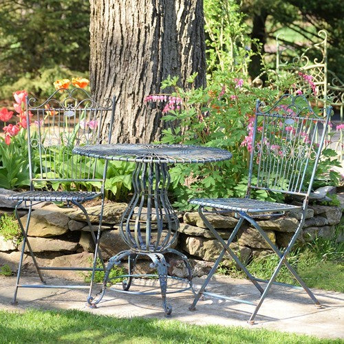 A three-piece outdoor bistro set featuring a round metal table and two matching folding chairs with a distressed blue-gray finish, set on a stone patio in a garden with a large tree trunk behind it.