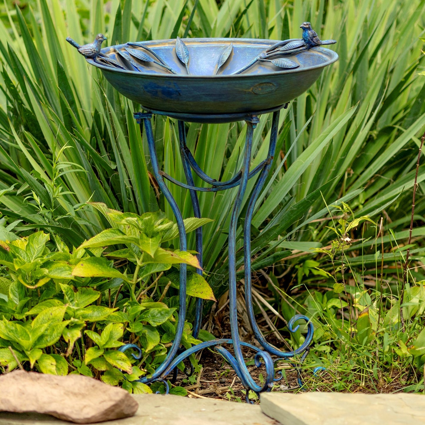 A decorative outdoor bird bath and stand set in a garden, featuring a blue metal basin with bird perched figures around the rim and a matching blue ornate stand, surrounded by green foliage.
