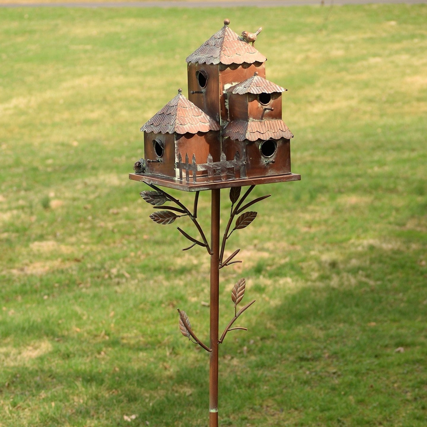 A rustic metal birdhouse garden stake with three small birdhouses mounted on a decorative branch-like stand, photographed outdoors on a grassy lawn.
