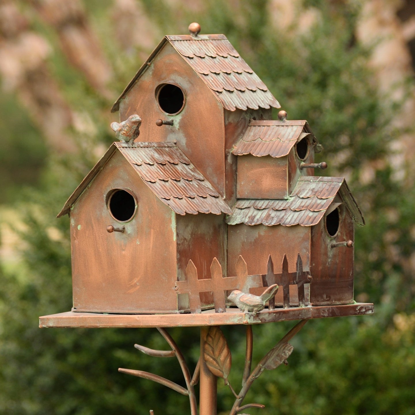 A rustic metal birdhouse set designed like small cottage roofs with a weathered copper and green patina finish, mounted on a decorative metal stand against a blurred outdoor garden background.