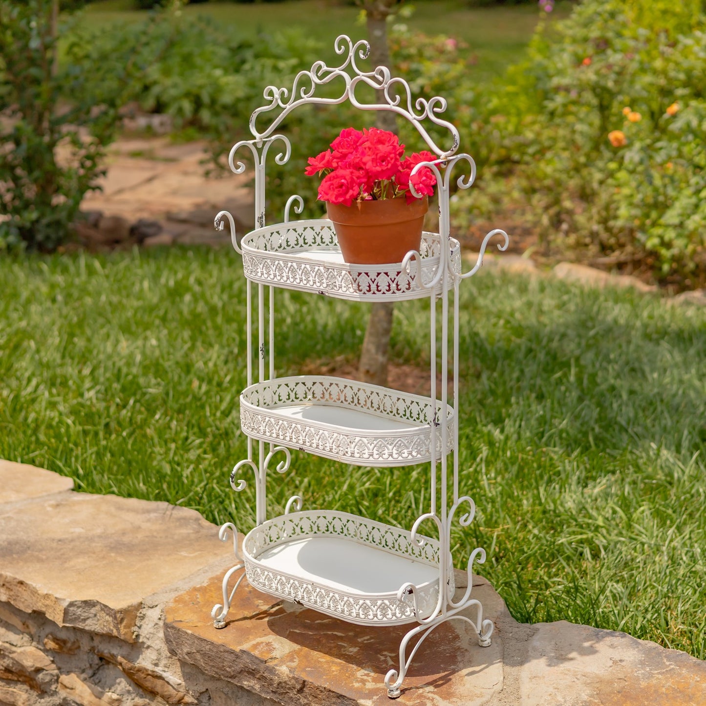 A three-tier white metal garden canteen stand with ornate scrollwork, displayed outdoors on a stone ledge with green grass and plants in the background. A potted pink flower sits on the top shelf.