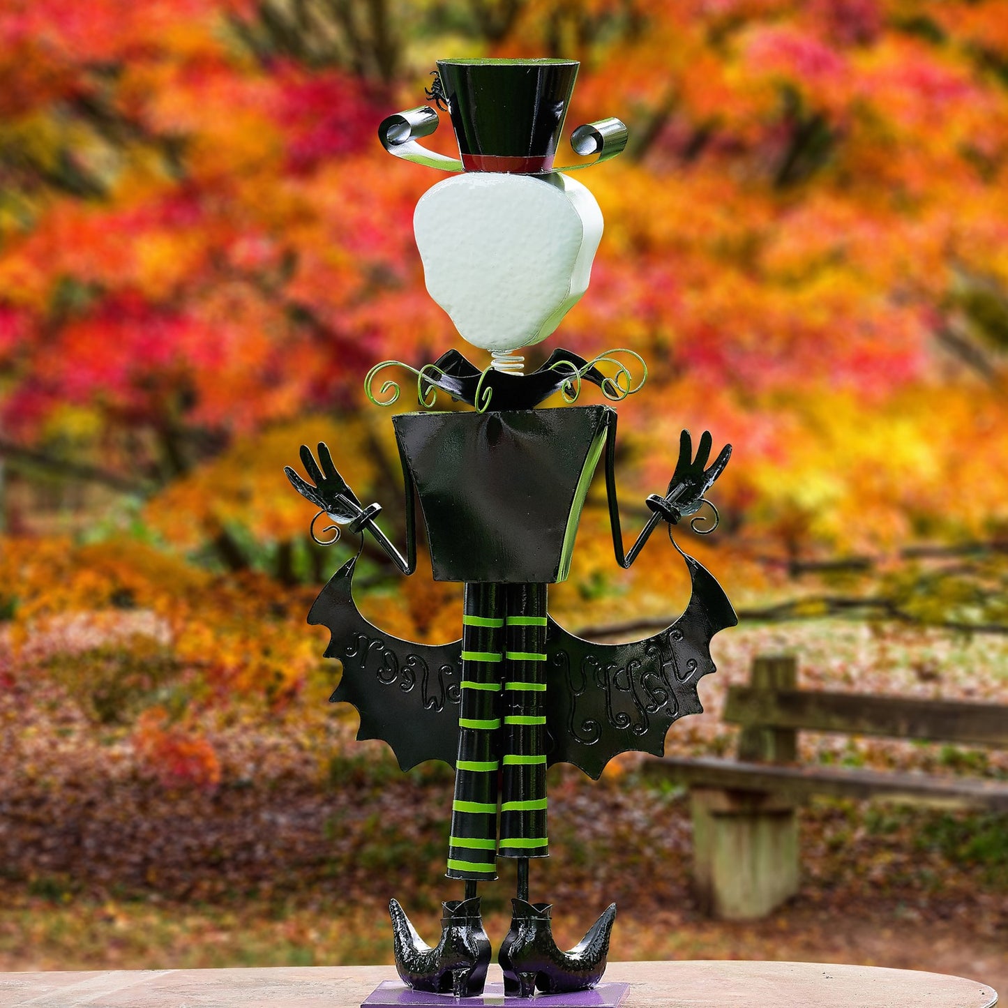 A Halloween metal garden statue of a stylized figure with a white head, black top hat, bat wings, and green-and-black striped legs, displayed outdoors against a blurred autumn foliage background.