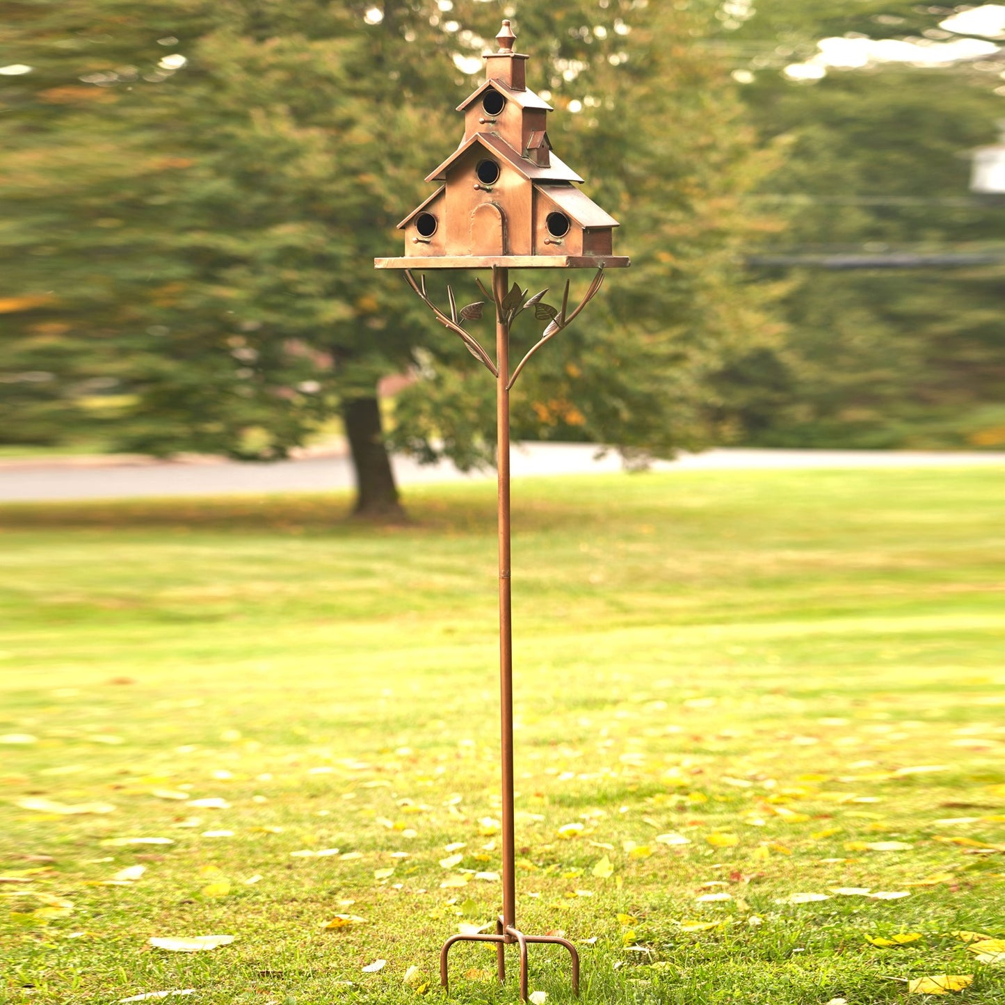 A rustic metal garden stake holding a multi-tiered birdhouse with a cottage-style roof and small bird perches, photographed outdoors on a grassy lawn with blurred trees in the background.