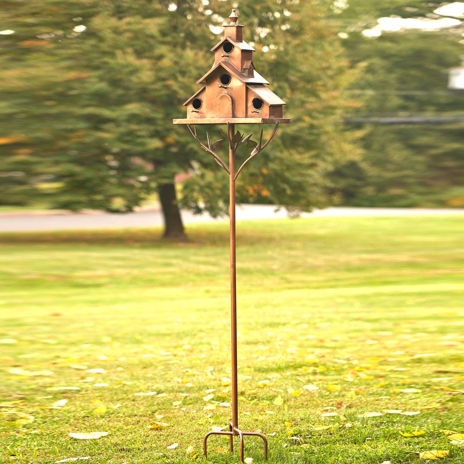 A rustic metal garden stake holding a multi-tiered birdhouse with a cottage-style roof and small bird perches, photographed outdoors on a grassy lawn with blurred trees in the background.