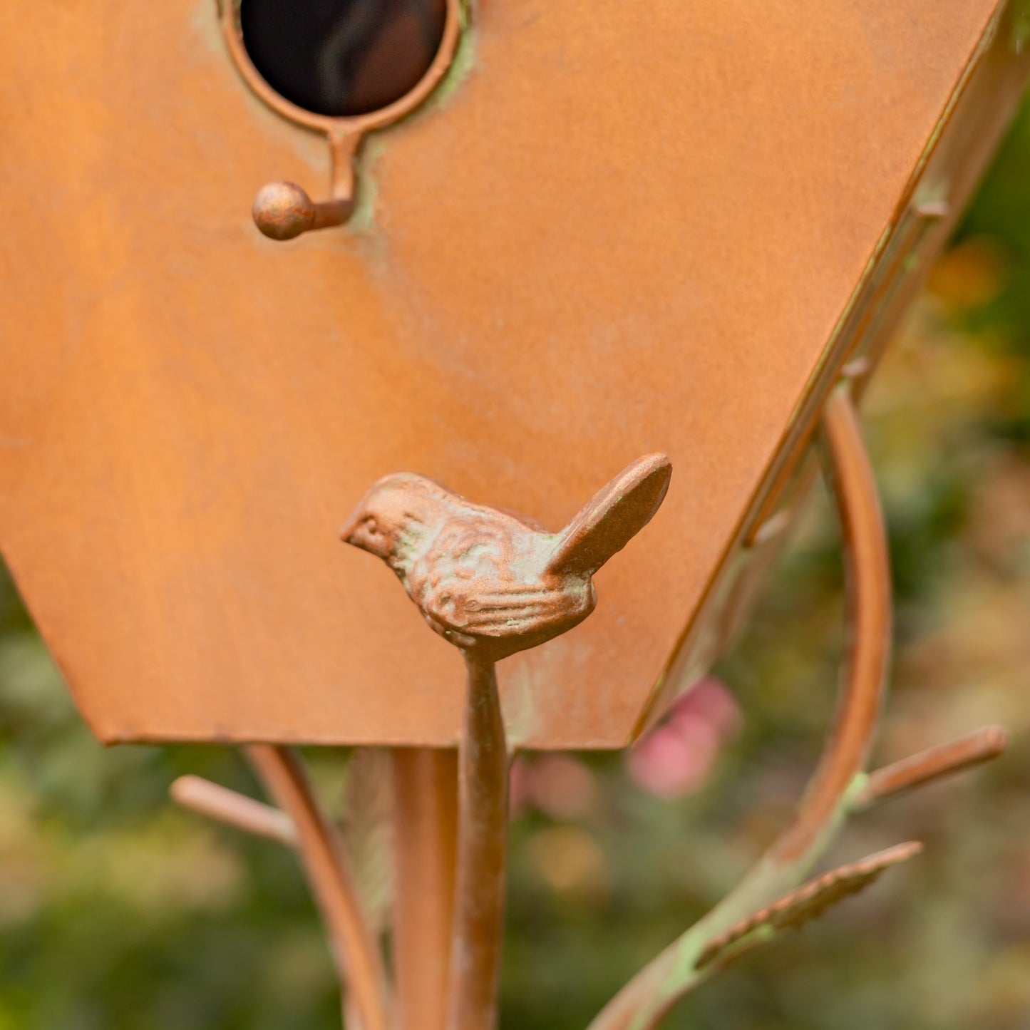 Decorative copper birdhouse with a small bird on top, set against a garden background with flowers and trees