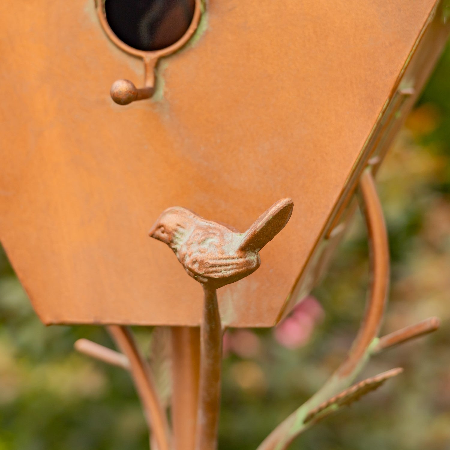 Decorative copper birdhouse with a small bird on top, set against a garden background with flowers and trees