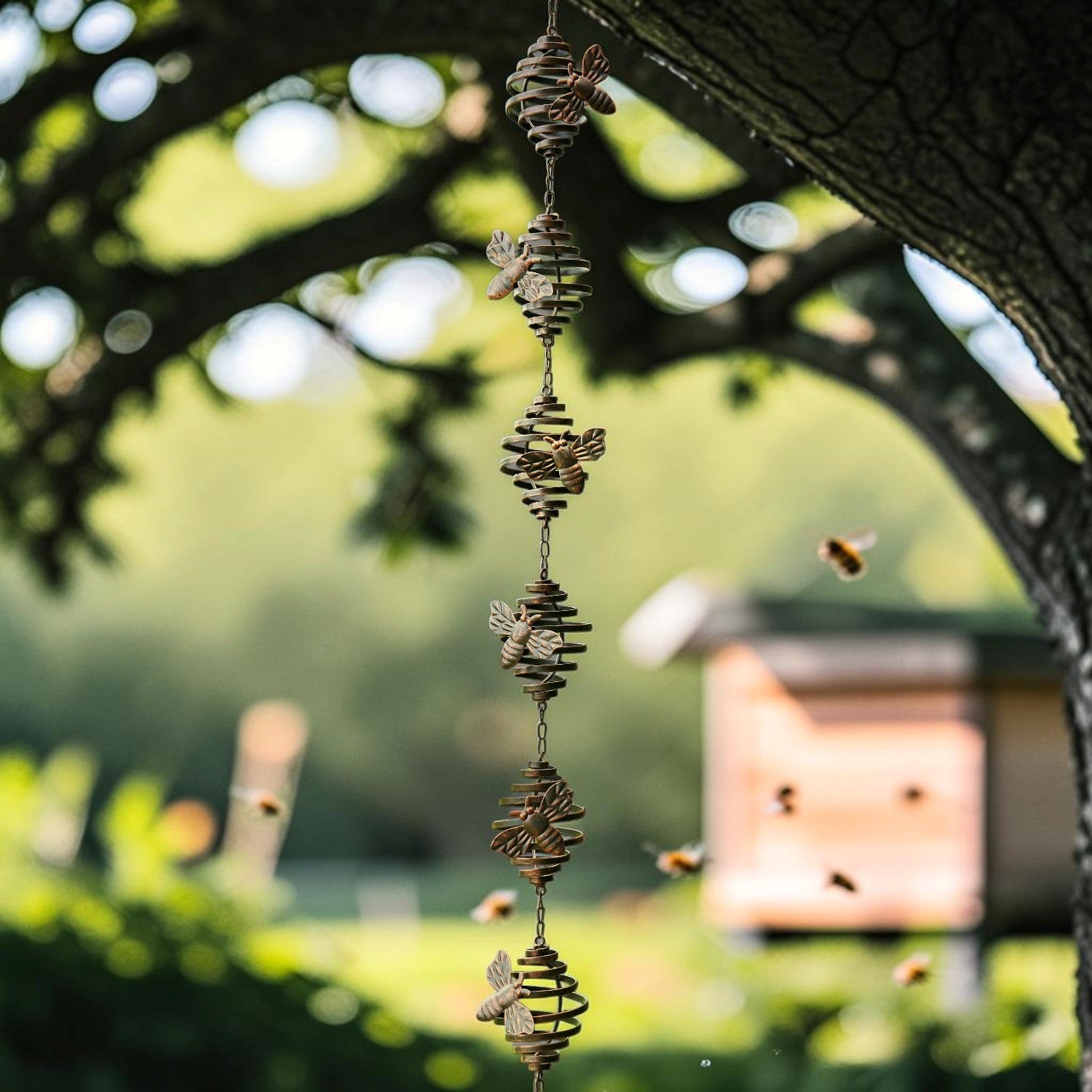 A hanging metal wind spinner with stacked insect and leaf-like shapes, photographed outdoors under a tree with bees flying nearby and a blurred garden background.