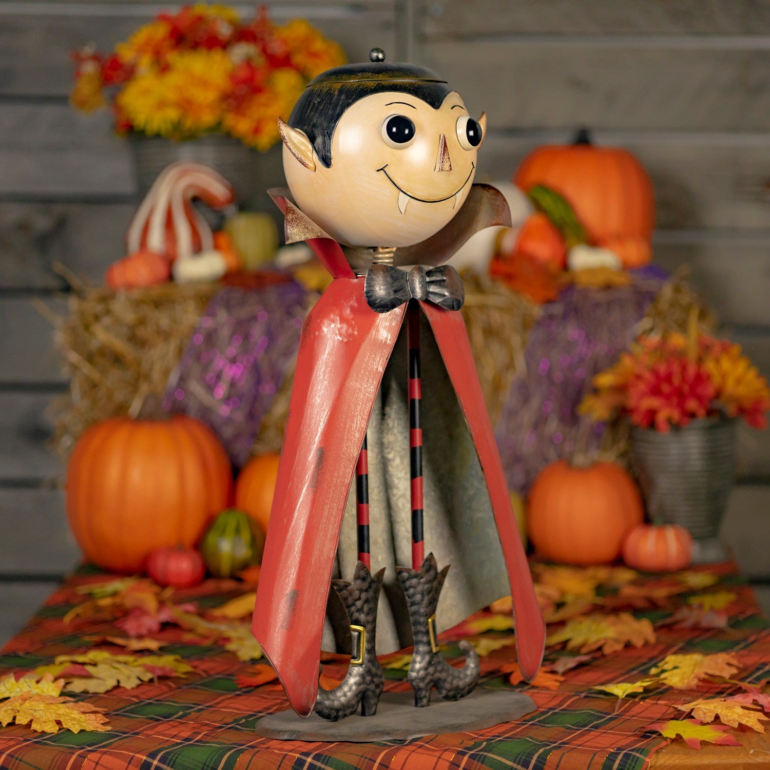 A Halloween decorative vampire figurine with a large round beige face, black hair, pointed ears, a black bow tie, and a red cape, standing on a plaid autumn tablecloth with pumpkins, hay bales, and fall flowers in the background.