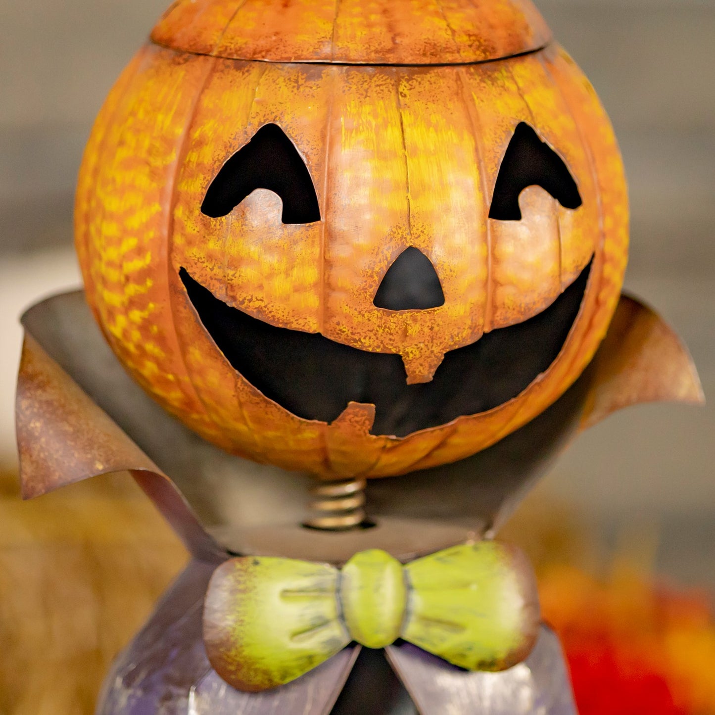 A Halloween jack-o'-lantern pumpkin decoration with a carved face, dark mouth, and a green bow tie, photographed close up against a softly blurred background.
