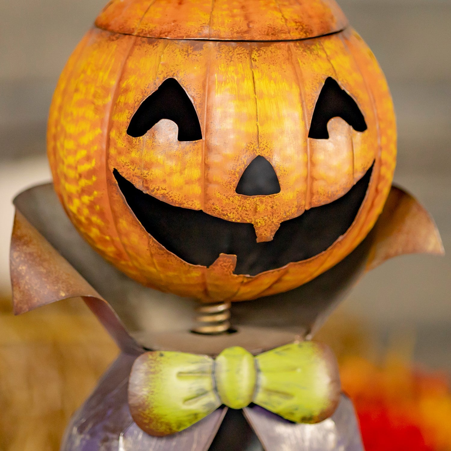 A Halloween jack-o'-lantern pumpkin decoration with a carved face, dark mouth, and a green bow tie, photographed close up against a softly blurred background.