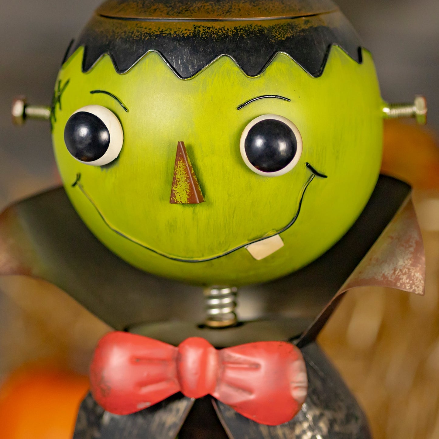 A close-up of a decorative Frankenstein-style pumpkin bobblehead with a bright yellow-green head, black hair, large black-and-white eyes, a small red nose, stitched smile, and a red bow tie on a dark garment.