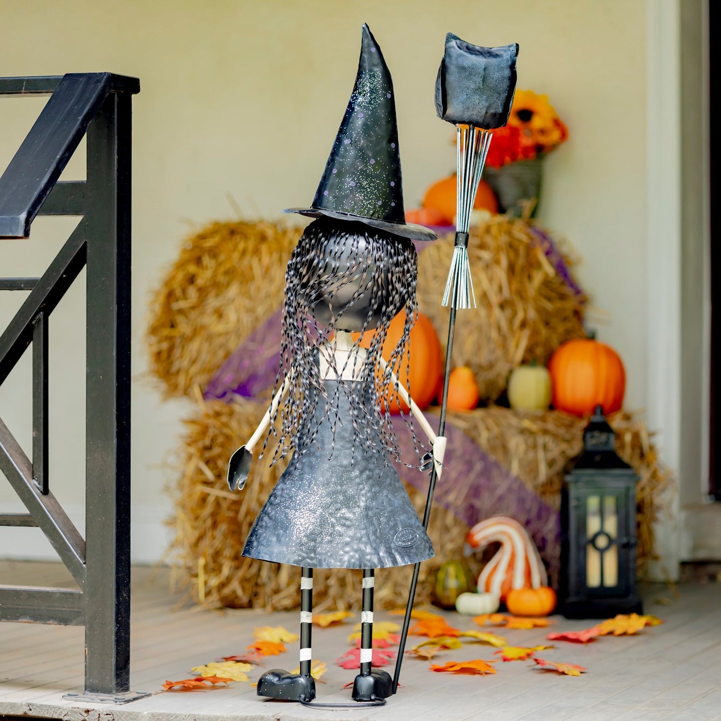 A decorative witch figurine stands on a porch in front of stacked hay bales and pumpkins. The figurine wears a tall black pointed hat and a silver glittery dress, with long black-and-white striped legs and holds a black staff topped with a gray square ornament.