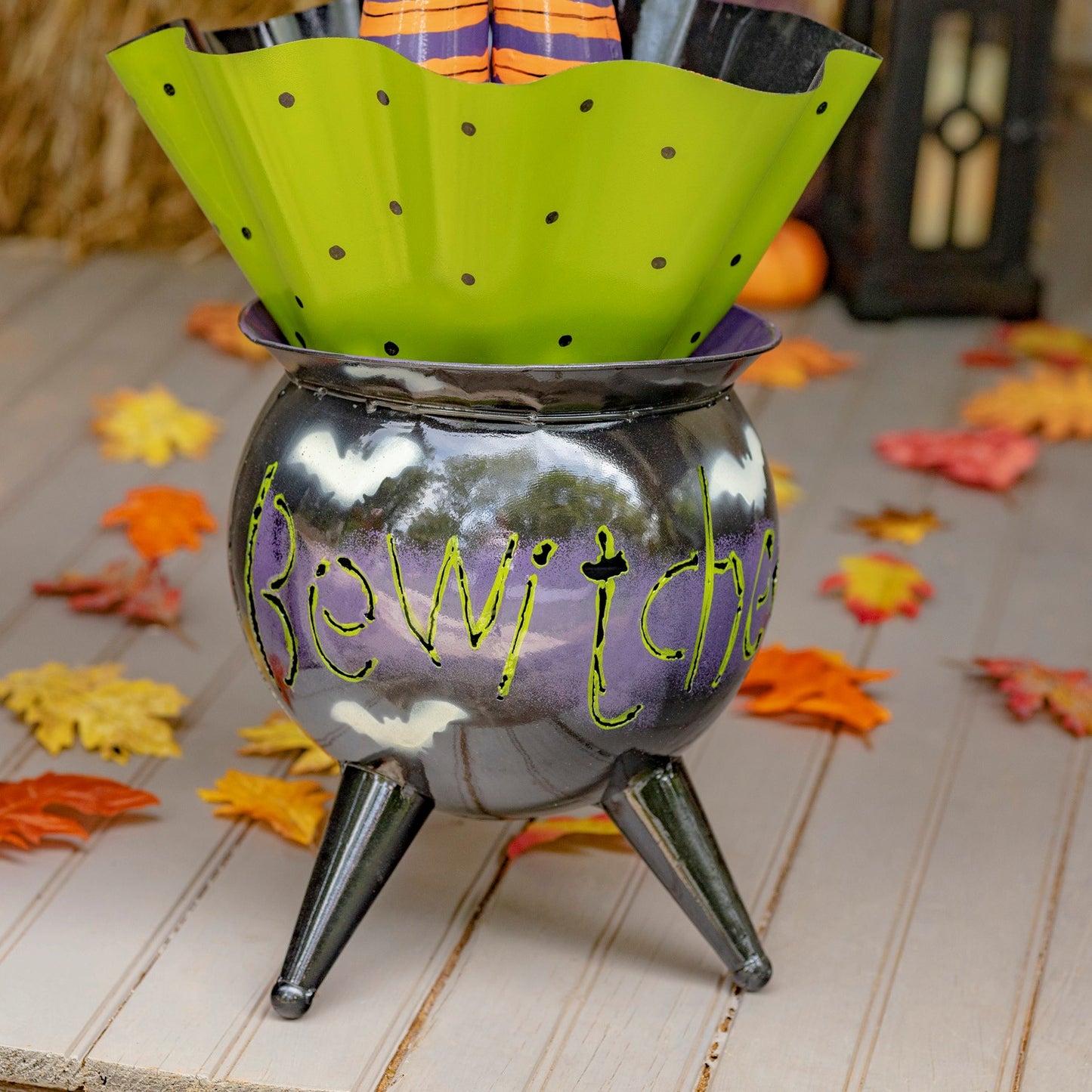 A Halloween-themed cauldron-shaped container with three legs, decorated with the word 'Beware' in green script over a purple and gray reflective surface. A bright lime green polka-dotted paper liner is draped over the top, with a striped candy bag peeking out.